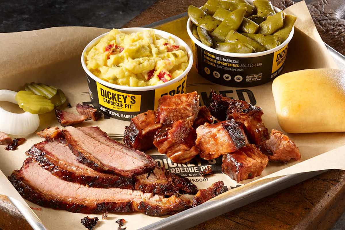 A close-up of sliced brisket, burnt ends, sides of mashed potatoes and green beans, with pickles and onions on a tray.